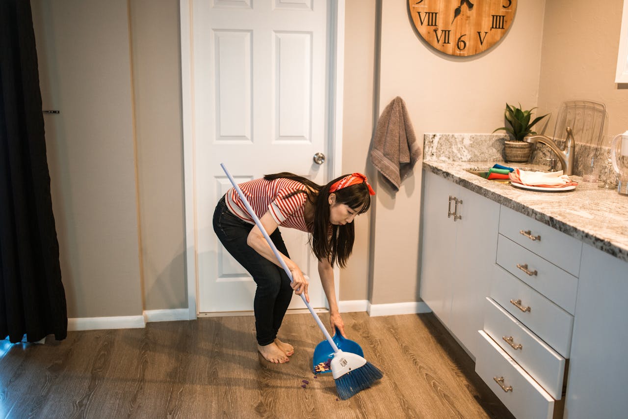 Asian woman sweeping kitchen floor, performing a household chore. Modern kitchen interior with granite countertop.