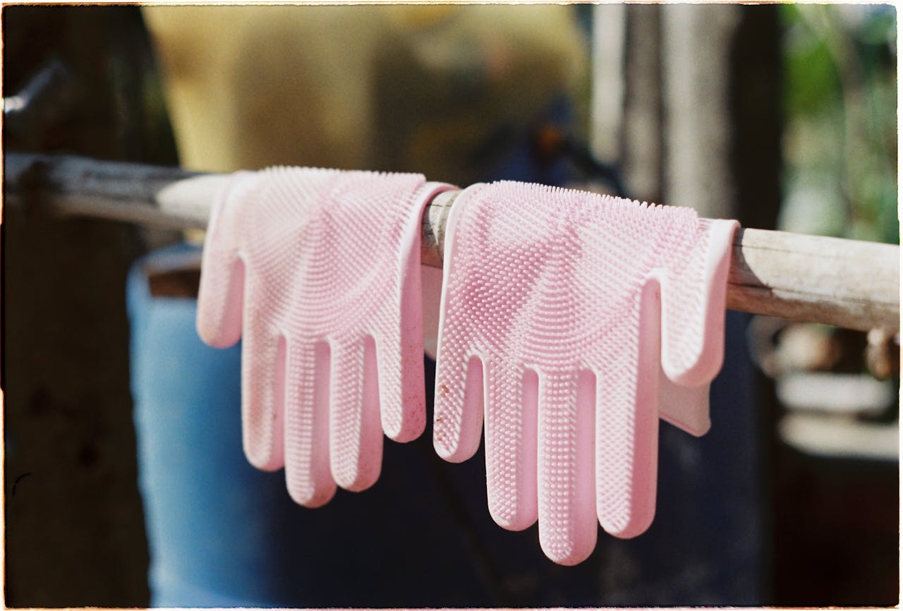 heros-img Two pink rubber cleaning gloves hanging to dry on a wooden stick outdoors in sunlight.