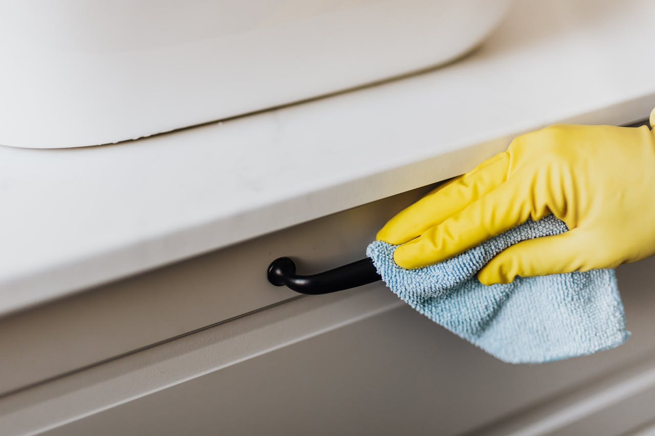 about-us From above of crop faceless person in yellow gloves cleaning handle of cabinet with microfiber cloth in modern bright bathroom